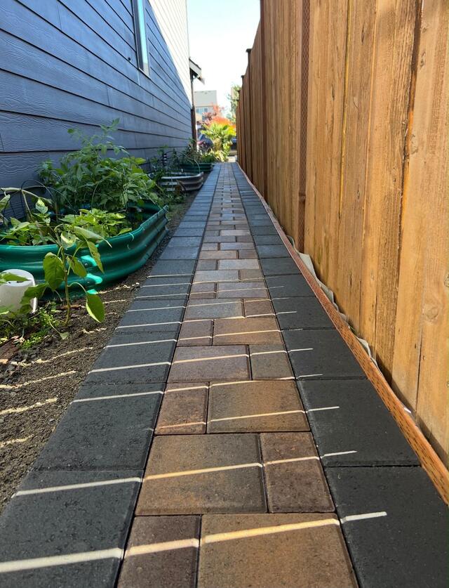 Paver walkway beside raised garden beds and a cedar fence
